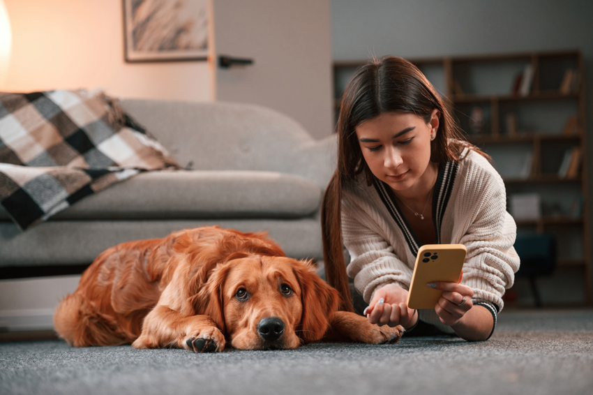 A dog and a teenage girl laying on the floor, girl is looking at a phone