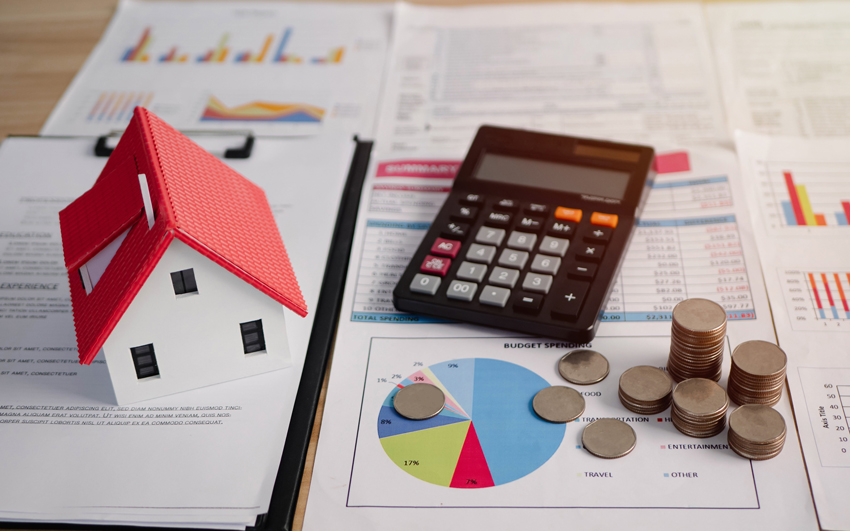A toy house, a calculator, and stacks of coins sitting on documents with charts and graphs
