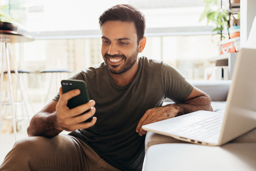 Man sitting on the floor holding a phone next to a sofa with a laptop on it